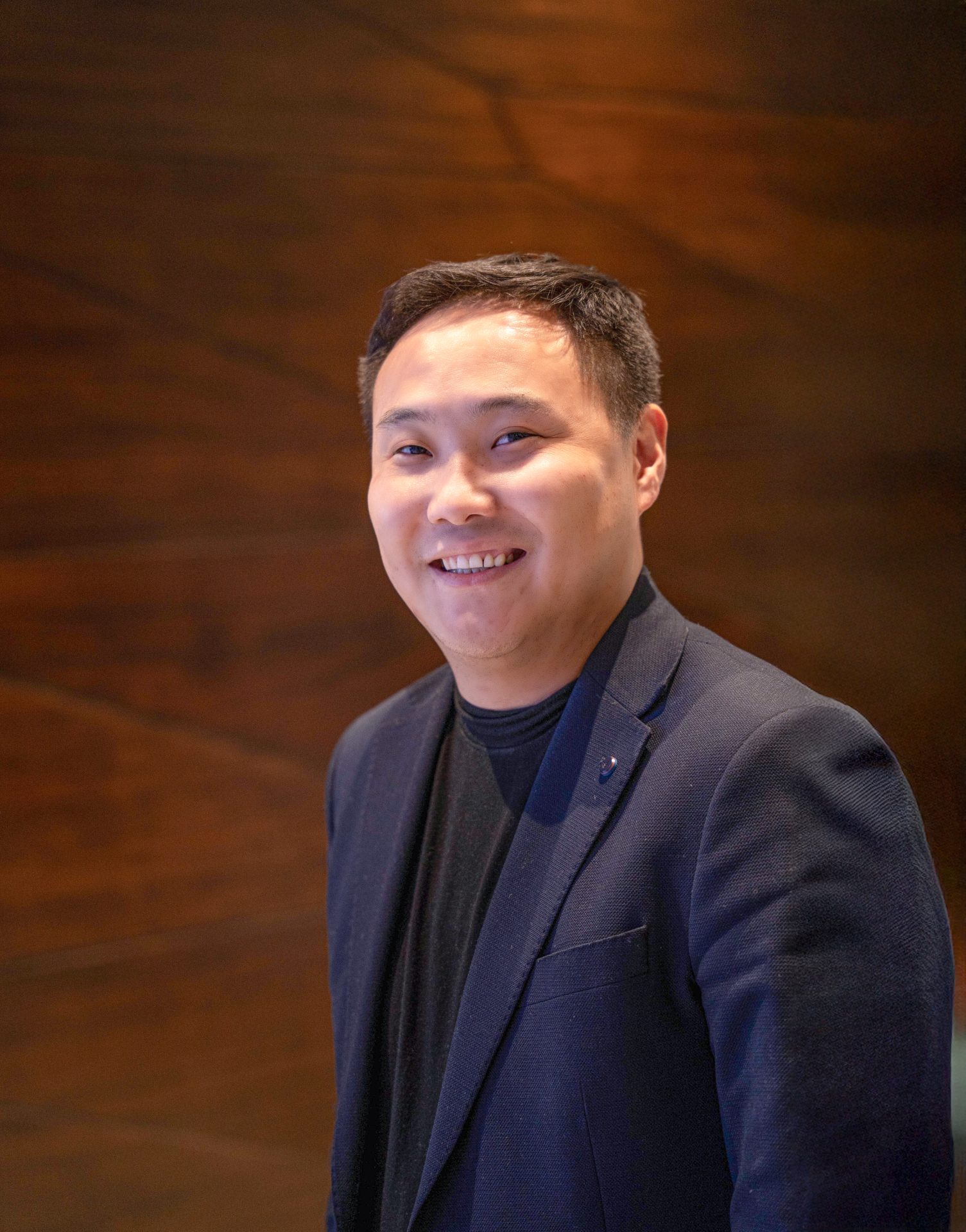 A smiling man in a dark blazer and black shirt stands against a blurred brown background.