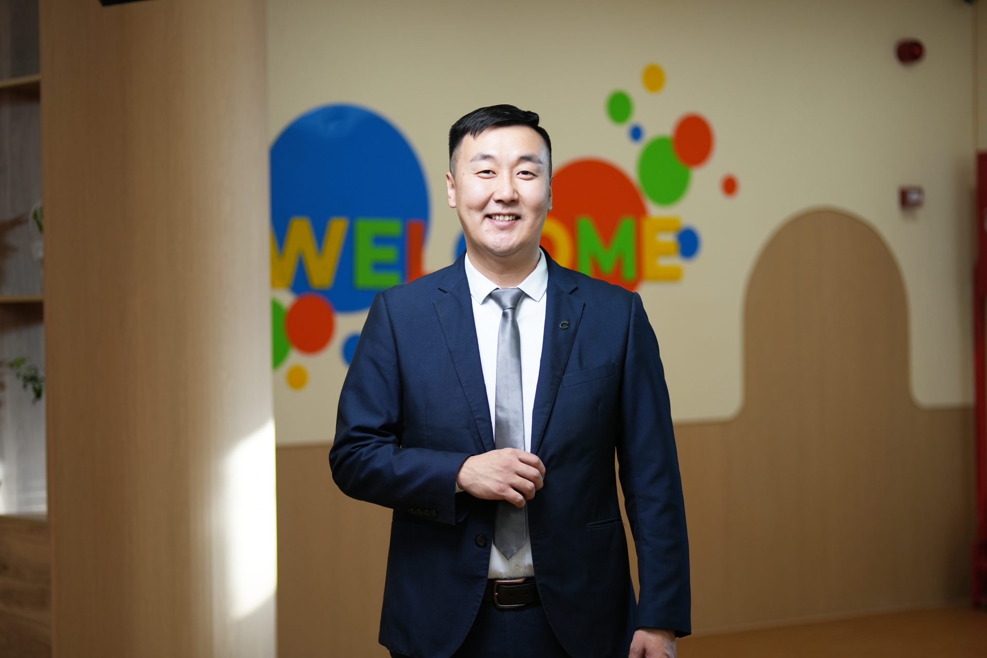 A man in a suit stands smiling in a room with colorful "WELCOME" letters on the wall behind him. The room has light wood and a cheerful atmosphere.