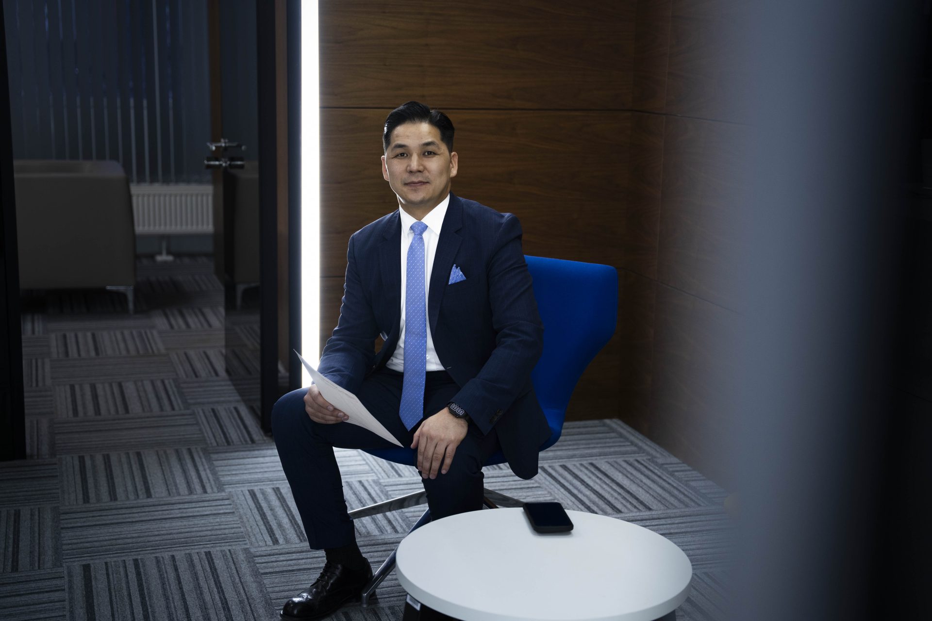 A man in a suit sits on a blue chair in an office, holding papers and looking at the camera. A smartphone is placed on a small round table in front of him. The room has a modern design with dark walls and carpeted flooring.