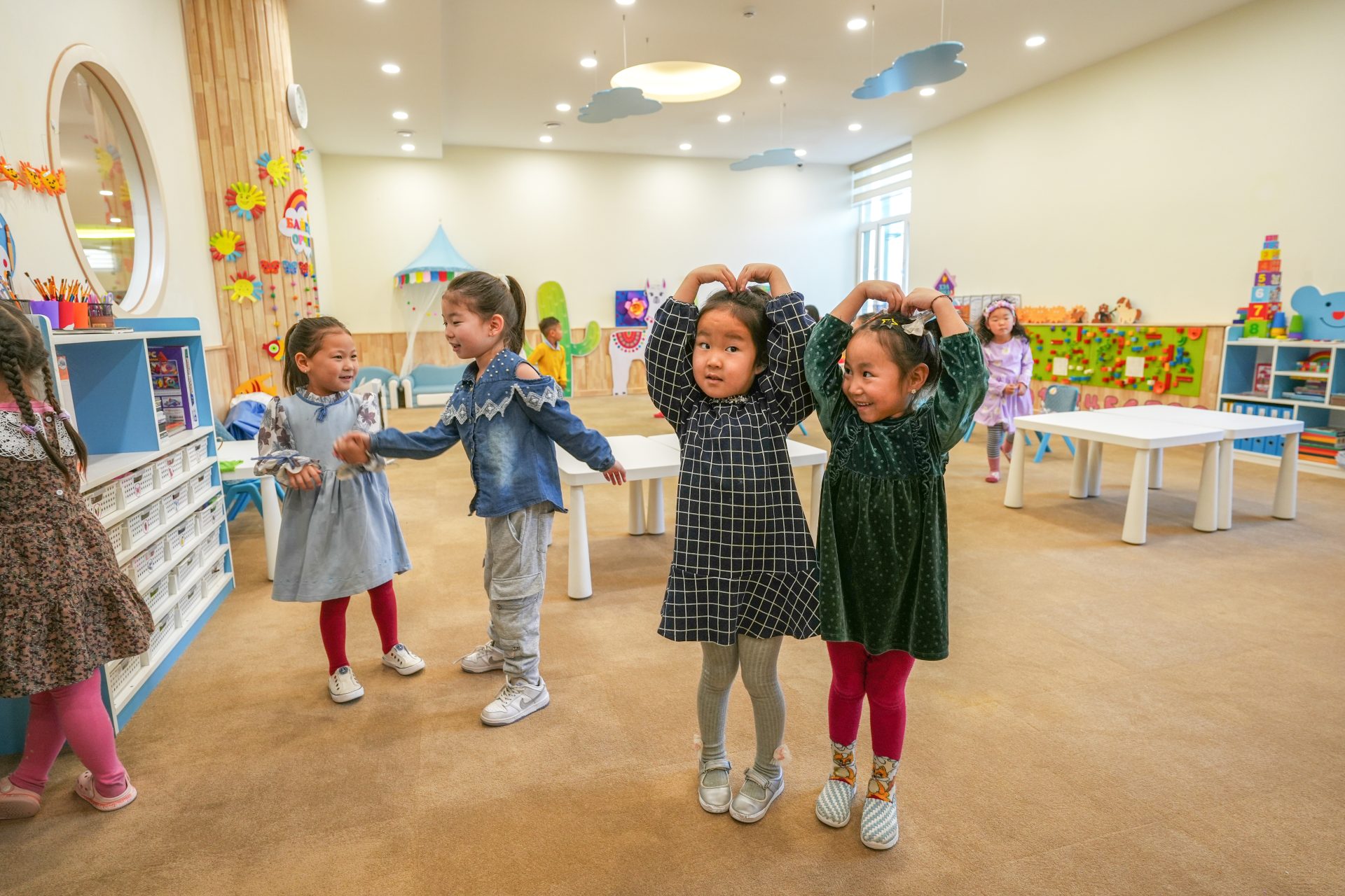 Children play together in a colorful classroom with whimsical decorations. Two girls in the center make heart shapes with their arms. The room is filled with tables and playful elements, creating a joyful learning environment.