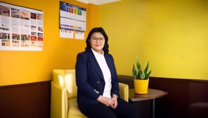 A person with long dark hair and glasses sits on a yellow chair in a room with bright yellow walls. There's a potted plant on a table beside them and various posters on the walls. They wear a navy blazer and white shirt.