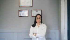 A person with long dark hair and glasses stands with arms crossed, wearing a white shirt. Behind them, four certificates or diplomas are framed and mounted on a gray wall. The setting appears to be a professional office.