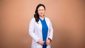 A person with long hair and glasses is wearing a white lab coat over a blue outfit, standing confidently against a plain brown background.