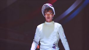 A fencer wearing a white fencing uniform and helmet looks forward with a focused expression. The helmet is tilted slightly back, revealing the competitor's face. The background is dark, highlighting the athlete.