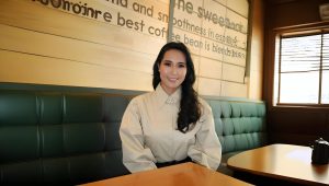 A person with long dark hair is sitting at a table in a cafe with wooden decor and green cushioned seats. They are wearing a light-colored blouse and smiling. The wall behind has a partially visible coffee-related sign.