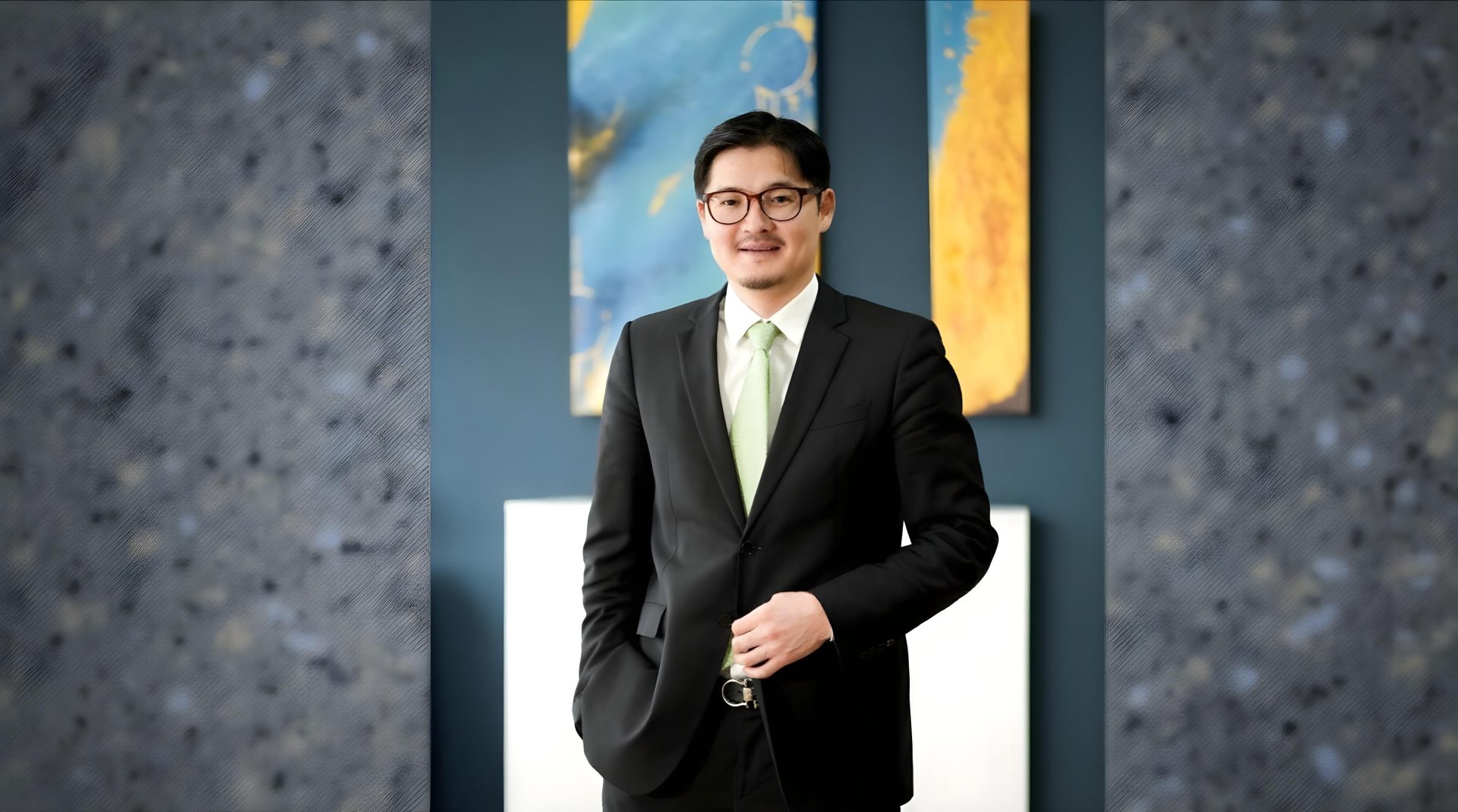 A man in a black suit and light green tie stands confidently in a room. The background features abstract artwork with blue and yellow hues.