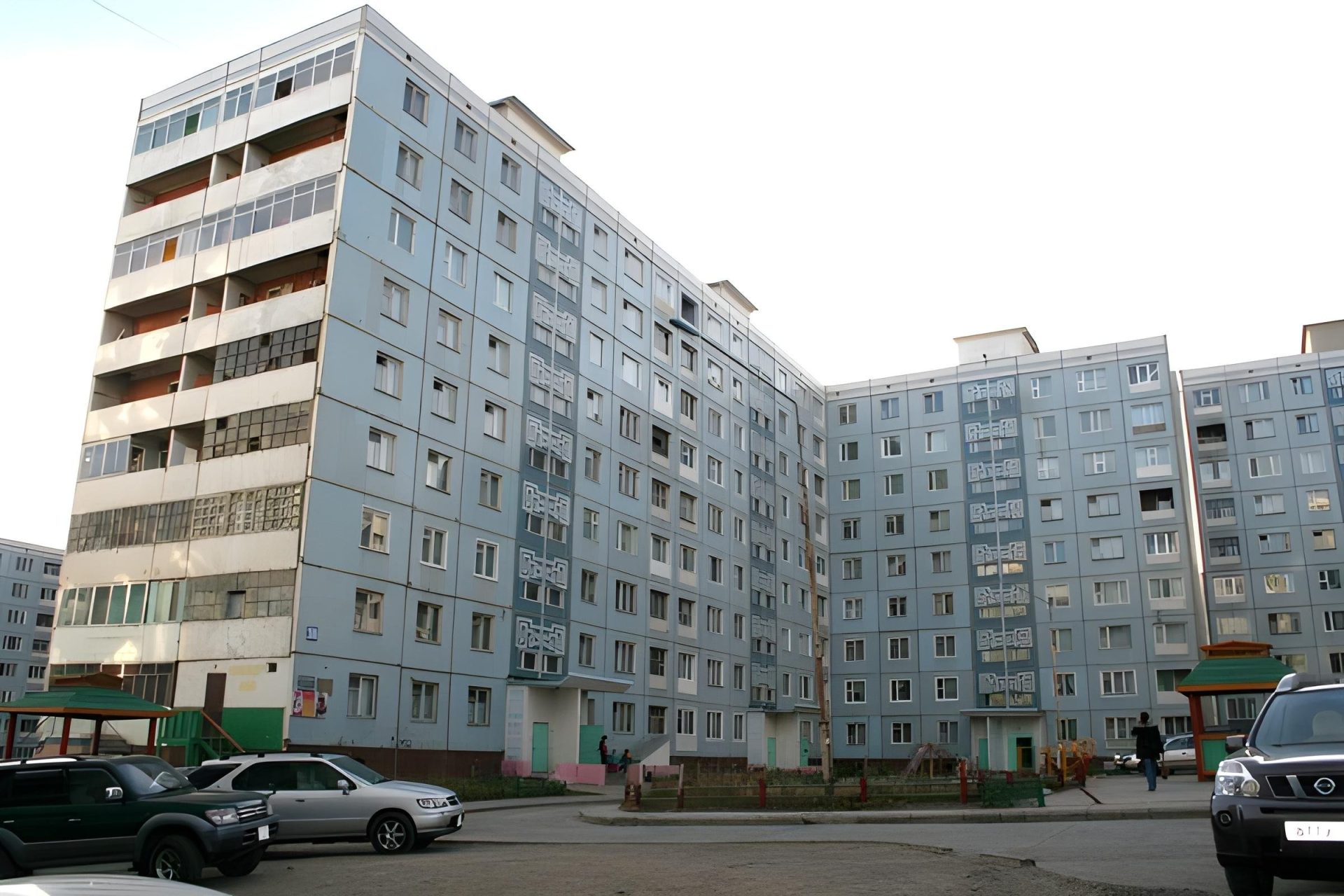 A large apartment building with a grey facade stands under a bright sky, reflecting last month's ups and downs. Several cars are parked in front. The building boasts multiple stories and numerous windows, with some balconies visible.