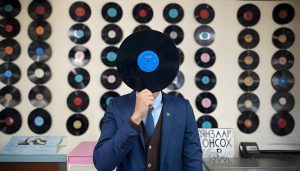 A person in a blue suit holds a vinyl record in front of their face, obscuring it. The background features a wall decorated with numerous vinyl records arranged in neat rows. A table with items is in the foreground.