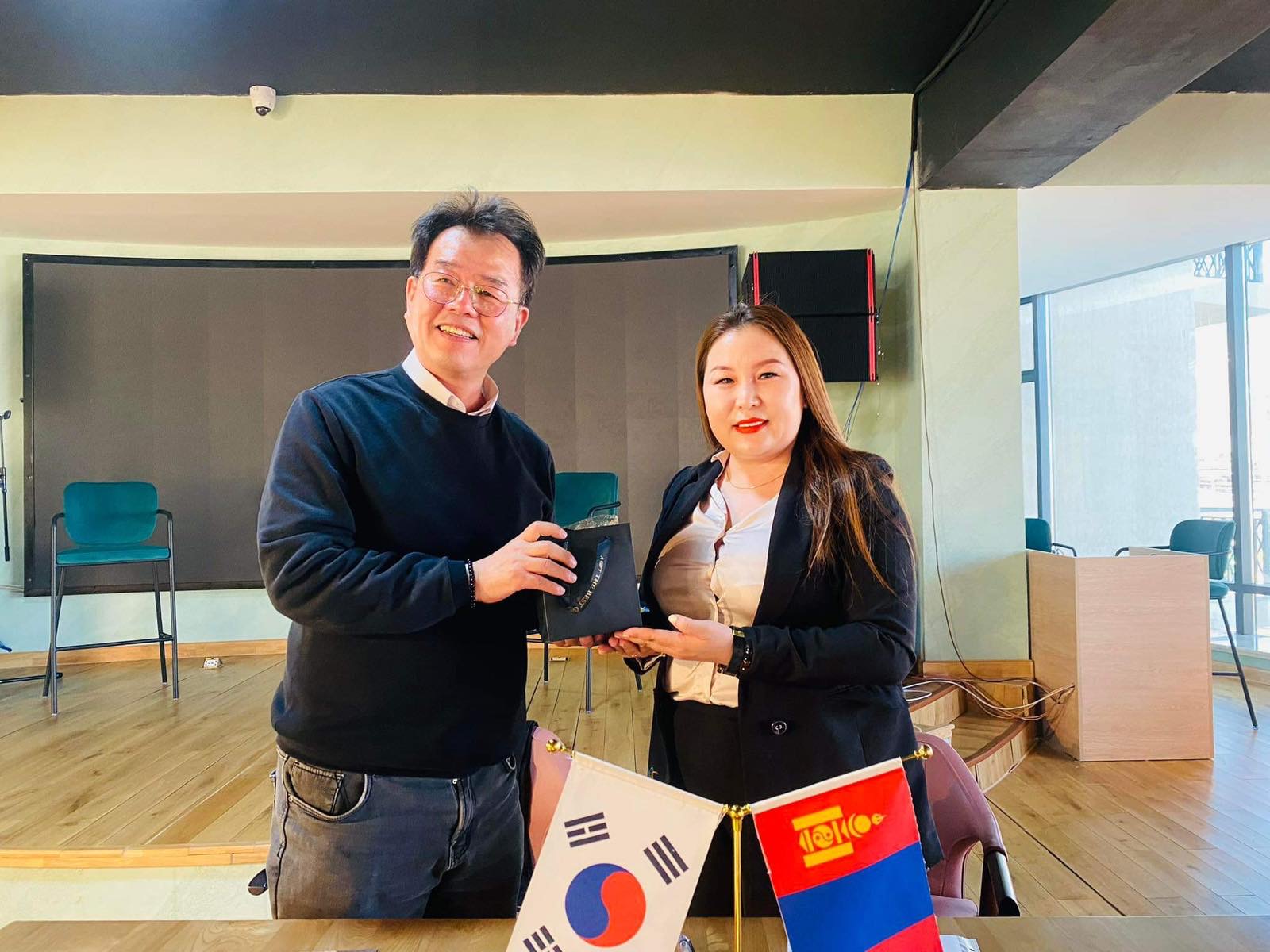 Two individuals stand in a room, exchanging a small black item. A table with South Korean and another flag is in front of them. The background features chairs and a presentation area with a screen. Both appear to be smiling.