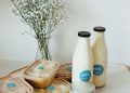 A selection of dairy products on a table, including bottles of milk, containers of cream, and a jar of yogurt. A bouquet of white flowers in a clear vase is in the background against a plain wall.