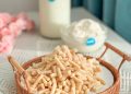 A woven basket is filled with crunchy, stick-like snacks on a table. In the background, there's a bottle of milk and a bowl of cream, both labeled with a blue sticker. Pink flowers are visible in the top left corner, adding a decorative touch.