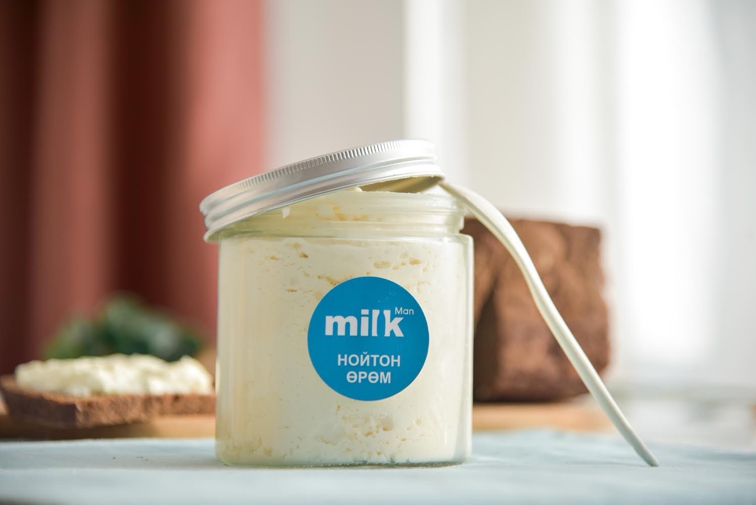 A jar of creamy substance with a partially open lid, featuring a blue label that says "milk" in white text and additional text in a non-Latin script. The jar is on a light surface with blurred objects in the background.