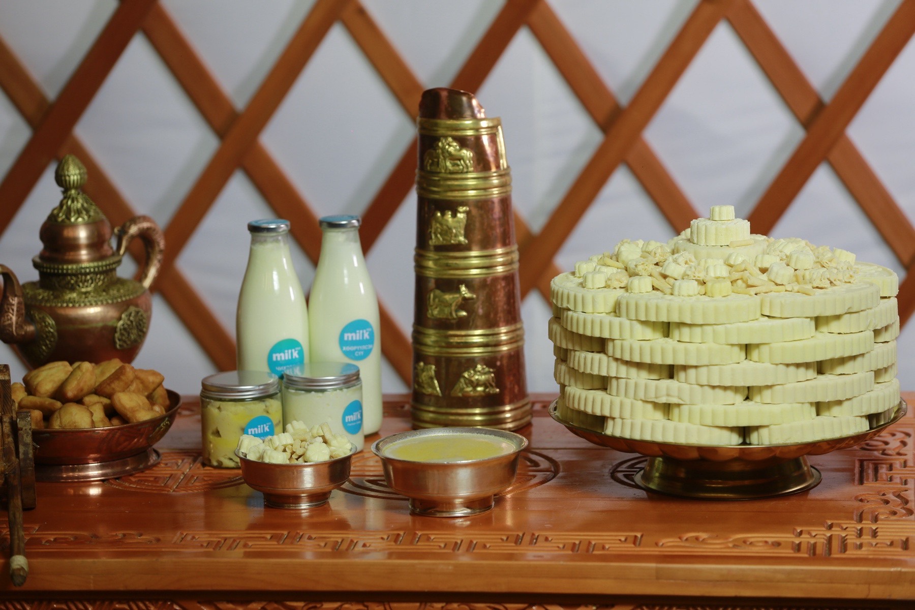 A traditional wooden table set with stacked white cheese, two milk bottles, a bowl of yellow liquid, metal pots, and intricate carvings. The background features a lattice pattern and ornate metalware, including a teapot.