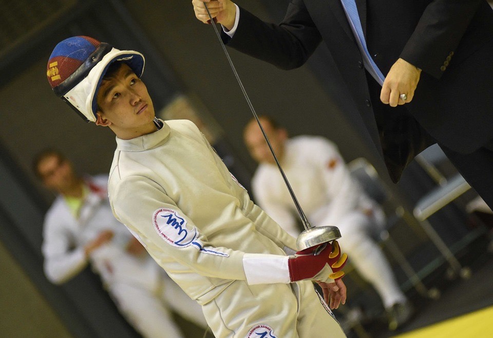 A fencer in a white protective suit and helmet holds a foil while standing next to a person in a suit, possibly a referee. The background shows blurred figures sitting, indicating a competition setting.