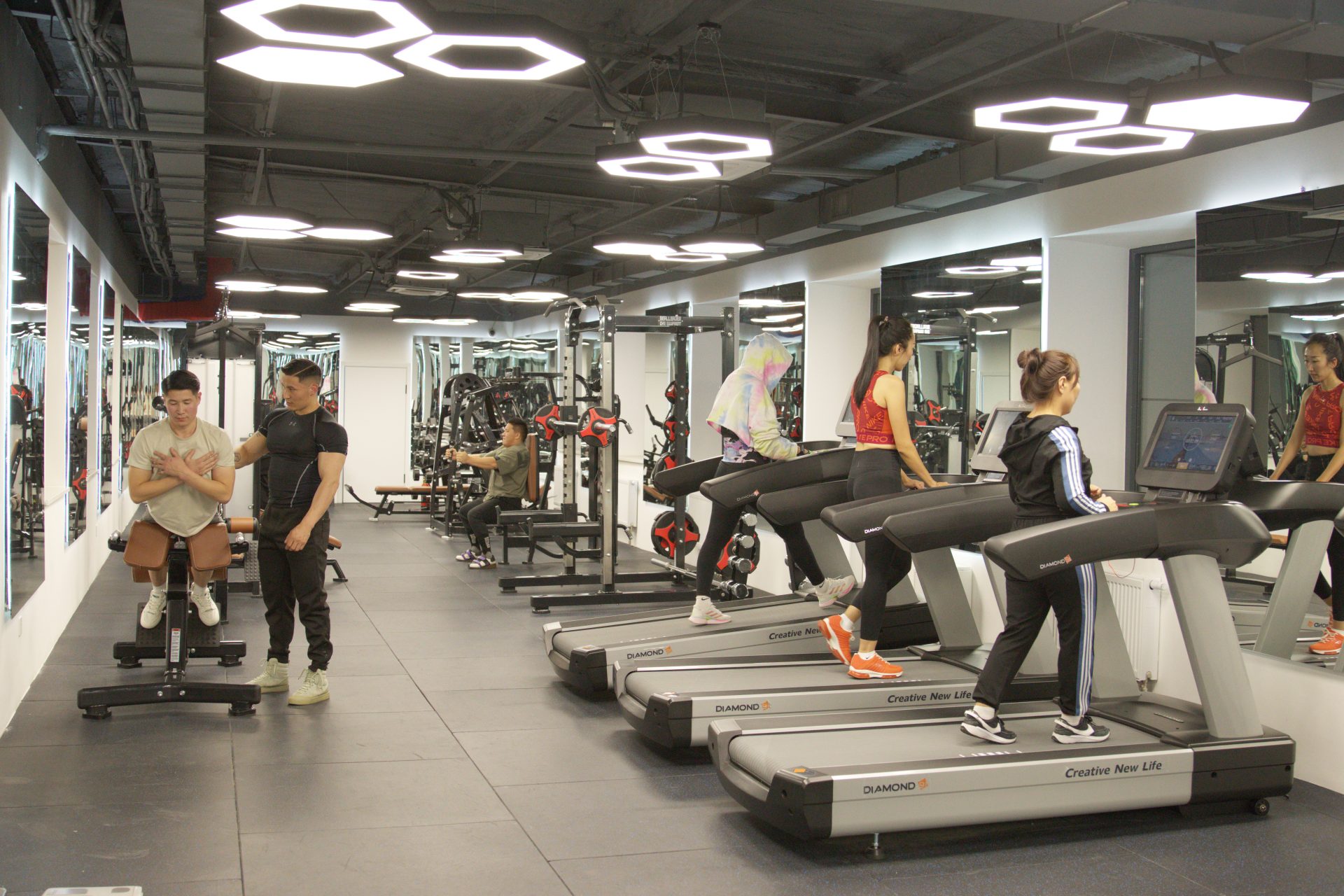 People exercising in a gym with treadmills and weight machines. Some are running on treadmills while others are using bench press machines. The gym is well-lit with modern ceiling lights and mirrored walls.