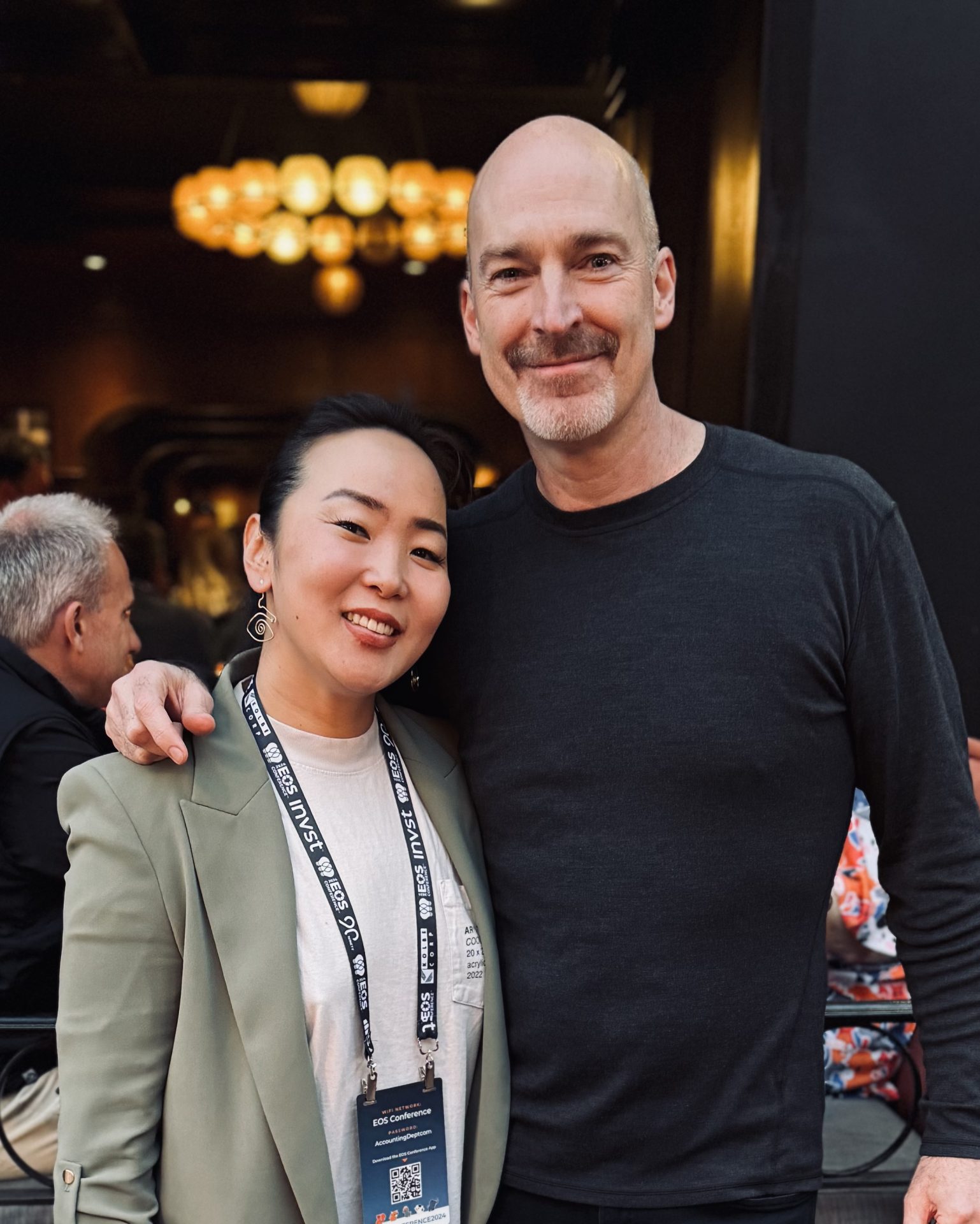 A woman and a man, embodying the spirit of Энтерпренер хүн, stand together, smiling at the camera. The woman wears a green blazer with a lanyard, while the man in a black long-sleeve shirt has his arm around her in a warmly lit indoor setting.