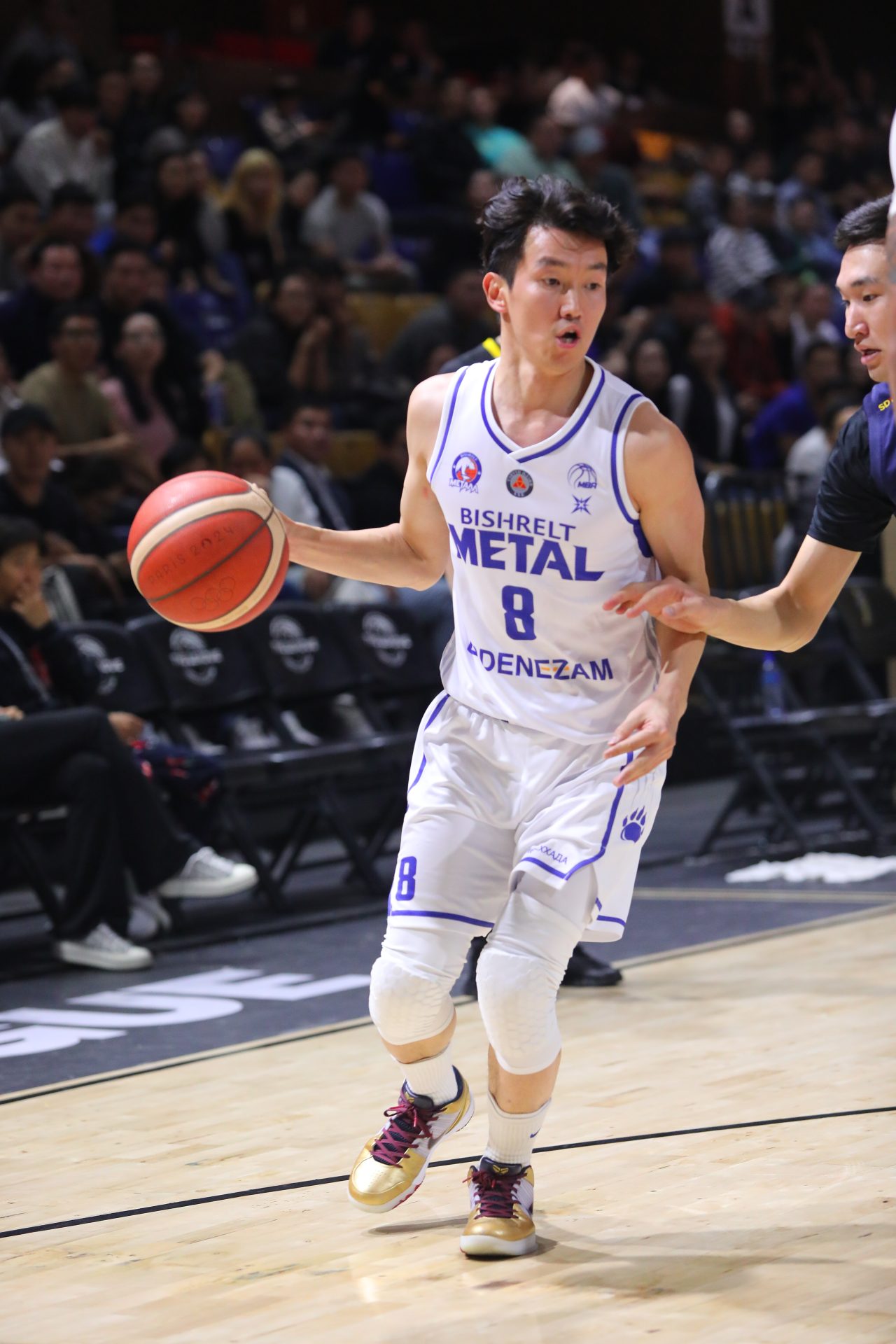 A basketball player in a white uniform, number 8, dribbles the ball on an indoor court. He is closely guarded by an opposing player wearing a dark uniform. The crowd is visible in the background.