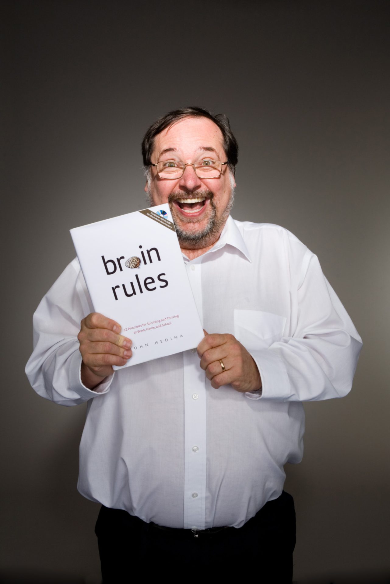 A man with glasses and a beard is smiling broadly while holding "Тархины дүрмүүд номын хураангуй." He is wearing a white shirt against a plain background.