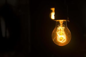 A glowing incandescent light bulb against a dark background, with a visible curlicue filament and a second bulb faintly illuminating in the distance.