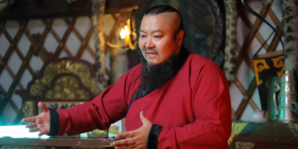 A man with a traditional hairstyle and a long beard is wearing a red robe. He is gesturing with his hands, appearing to speak. The background features wooden latticework and ornate decorations.