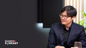 A man in a dark suit and glasses is seated, looking to the right, on a podcast set. The background features subtle lighting and a potted plant. The words "Business Podcast" are displayed on the left side of the image.