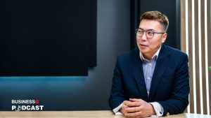 A person wearing glasses and a dark suit is sitting at a table, engaged in conversation. The background features a dark wall, and there's a "Business Podcast" logo in the corner.