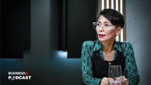 A person with short hair and glasses is sitting at a table, wearing a patterned shirt and dark vest. Engaged in conversation, they discuss "Энерги бардаг муу дадал зуршил хандлагуудаас хэрхэн салах вэ," with a blurred podcast logo in the corner and a lit background.