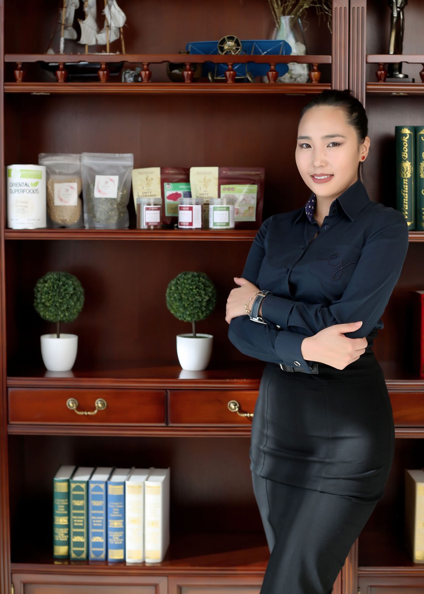 A woman in a dark blouse and skirt stands with arms crossed in front of a wooden bookshelf. The shelf holds decorative items, books, small round plants, and various packaged goods. She smiles slightly and looks at the camera.