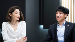 A man and a woman are seated at a table, engaged in conversation. The woman is wearing a white blouse and pearl necklace, while the man is in a dark suit with a white shirt. The background is softly lit with dark and light panels.