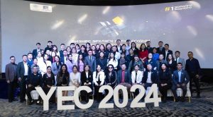 A large group of people, dressed in formal and casual attire, pose together on a stage with "YEC 2024" in large letters in front. A backdrop features geometric designs and text. The setting suggests a conference or event.