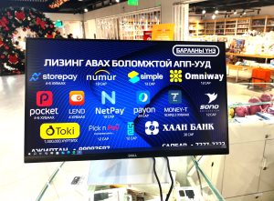 A computer monitor in a store displays various digital transaction logos with text in Cyrillic. Shoppers browse in the background, and shelves are filled with goods.