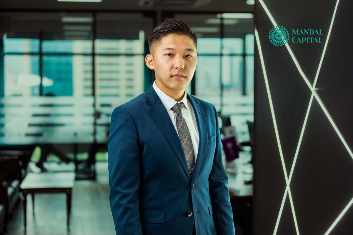 A man in a blue suit and striped tie stands confidently in an office with glass partitions and city views. The logo "Mandal Capital" is visible in the top right corner.