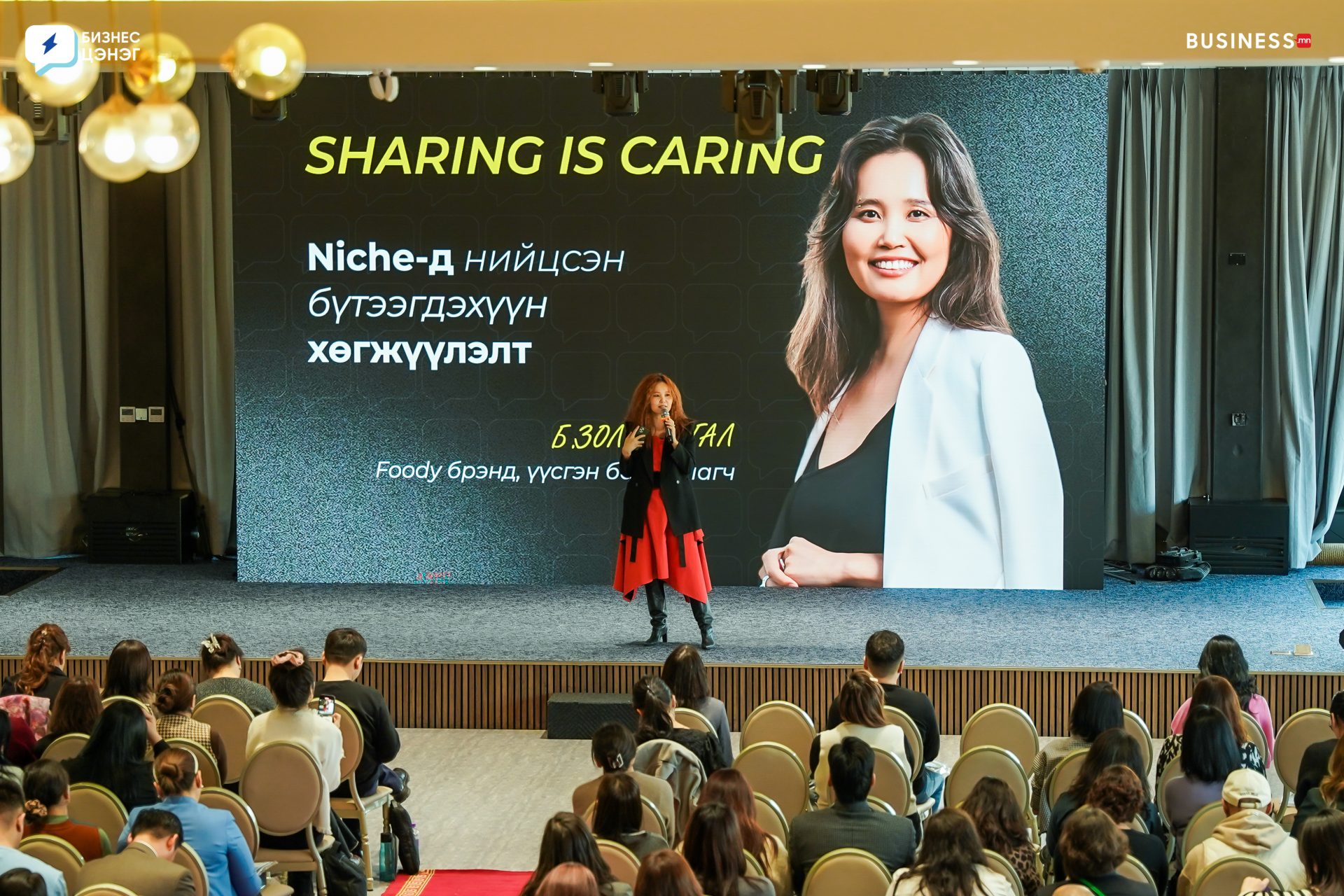 A woman in an orange dress and black jacket speaks on stage with a microphone. Behind her is a large screen displaying her image and the text "Sharing is Caring." An audience sits in rows facing the stage.