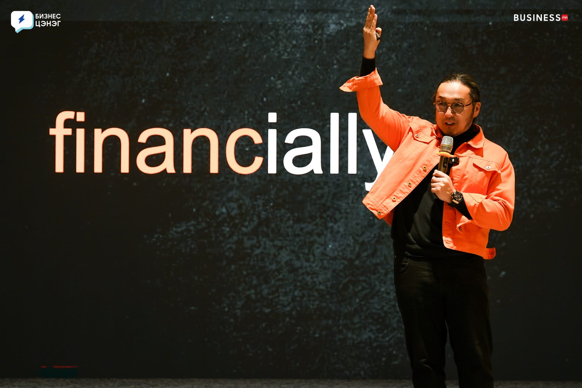 A person wearing a bright orange jacket and holding a microphone stands in front of a dark backdrop with the word "financially" visible. One hand is raised, as if making a point during a presentation or speech.