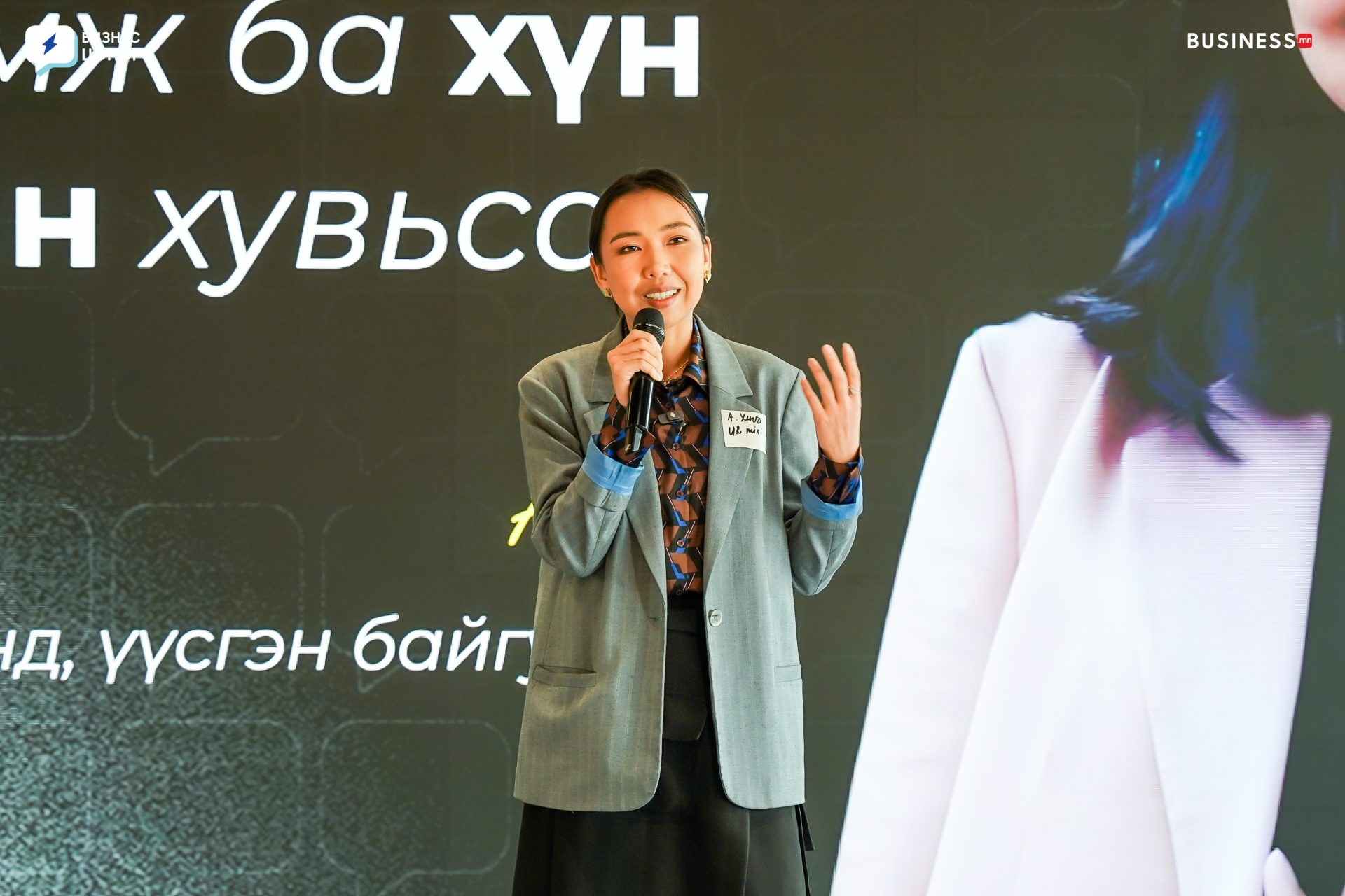 A person standing and speaking into a microphone, dressed in a blazer and patterned shirt. They have long hair tied back and are in front of a large screen with text. The background is part of a stage or presentation setting.