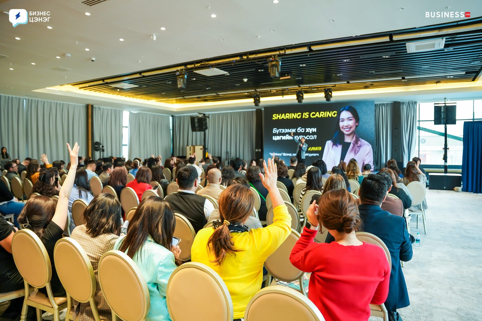 A conference room filled with seated attendees facing a large screen displaying a woman's image and the text "SHARING IS CARING." One person has their hand raised. The room has a modern design with ceiling lights and large windows.