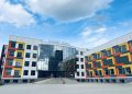 Modern building with a colorful facade featuring red, orange, and yellow geometric patterns. It has a central glass entrance with steps leading up to it. The sky above is partly cloudy.