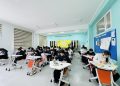 A classroom with students sitting at individual desks, focused on writing or working. The walls are painted teal, with a bulletin board and quotes. Large windows allow natural light to fill the room.