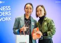 Two people stand side by side, smiling at the camera. One holds a book with an orange cover and gear designs. They are against a blue backdrop with the words "Business Builders Network.