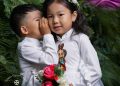 Two children dressed in white traditional outfits stand together. The boy is whispering to the girl, who is holding a bouquet of roses. She has a flower crown and an embroidered design on her dress. Green foliage is in the background.