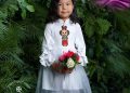 A young girl stands in a white dress with ruffled sleeves, holding a bouquet of pink and white flowers. She is in front of lush green foliage. The dress features a colorful embroidered design on the front. The background shows "Tumen Torgo" branding.