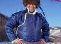 A person stands outdoors wearing a traditional blue patterned outfit and a fur hat. They are smiling with a snowy landscape and hills in the background, under a clear blue sky.