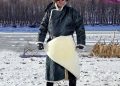 A person wearing traditional winter attire with a dark patterned coat, fur hat, and sunglasses stands on snow-covered ground near a partially frozen river, with barren trees in the background.