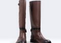 Brown leather equestrian boots with brogue detailing. One boot stands upright while the other leans slightly forward. Background is white with "Italian Design" text at the top left and a circular logo on the right.