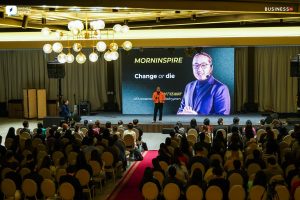 A speaker in an orange jacket passionately addresses the audience in a conference hall. Behind him, a large screen displays "MORNINSPIRE - Change or die" alongside his photo. Attendees listen intently, reminded that "Бизнесийн хамгийн үнэ цэнтэй зүйл нь та өөрөө.