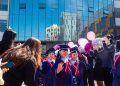 Children in blue graduation caps and gowns walk on a red carpet in front of a building labeled "Smart School." They are surrounded by people holding balloons and pom-poms, celebrating under a clear blue sky.