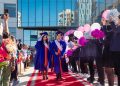 Two graduates in blue gowns walk down a red carpet lined with cheering people holding pom-poms. They smile as they approach a glass building adorned with balloons. A festive atmosphere surrounds the graduation ceremony.