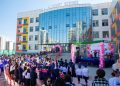 A graduation ceremony outside a modern school building with a glass facade. Graduates in blue gowns and caps sit while an audience watches. A stage is decorated with pink balloons, and a speaker addresses the crowd.