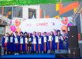 A group of graduates in blue and orange robes stand on a stage holding certificates. They are in front of a colorful backdrop with balloons and logos. The event is outdoors with a building in the background.