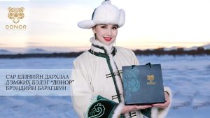 A person dressed in traditional Mongolian clothing smiles while holding a gift bag in a snowy landscape. The bag has "Donor" branding, and text in a non-English language is visible alongside a circular logo.